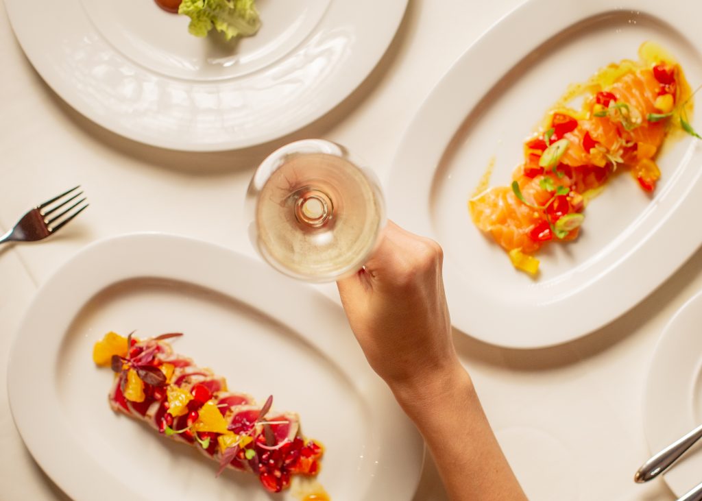 Image of a vibrant assortment of colourful foods displayed on plates on a table, with a hand holding a glass of wine above.