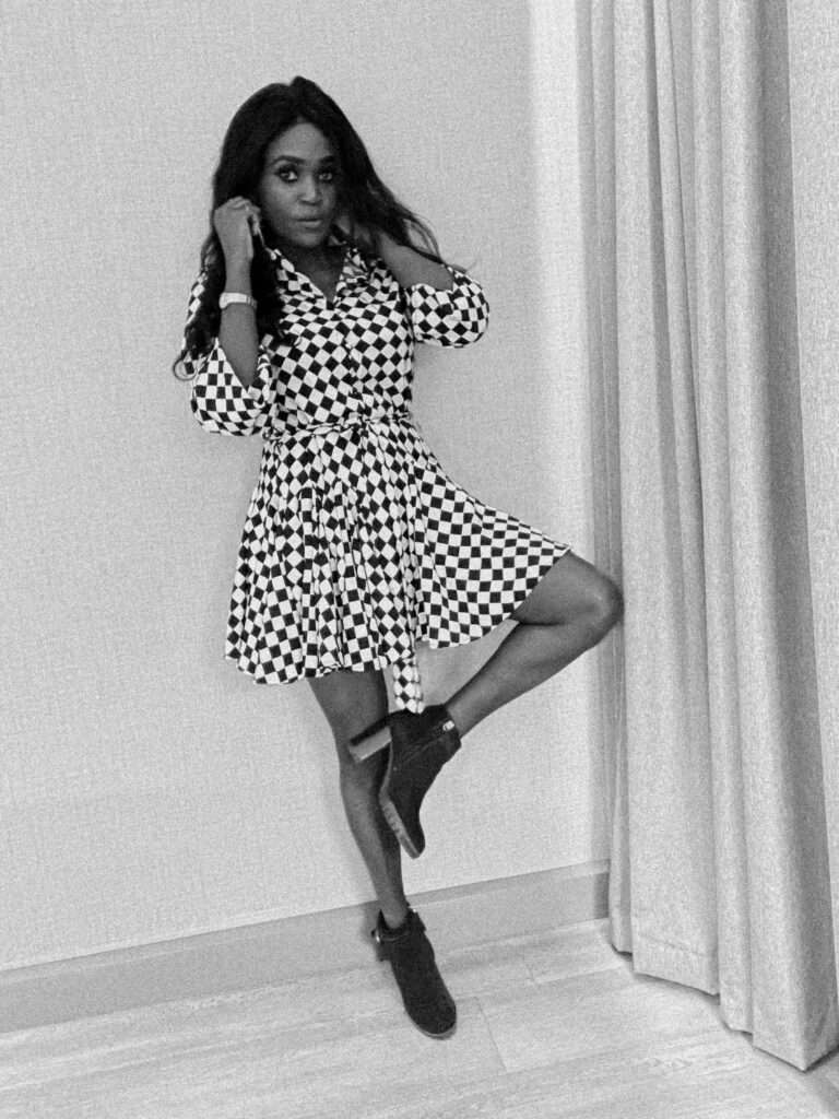 A woman standing indoors, posing with one leg slightly lifted. She is wearing a black-and-white checkered dress and black ankle boots, perfect to transition your summer outfits into fall.