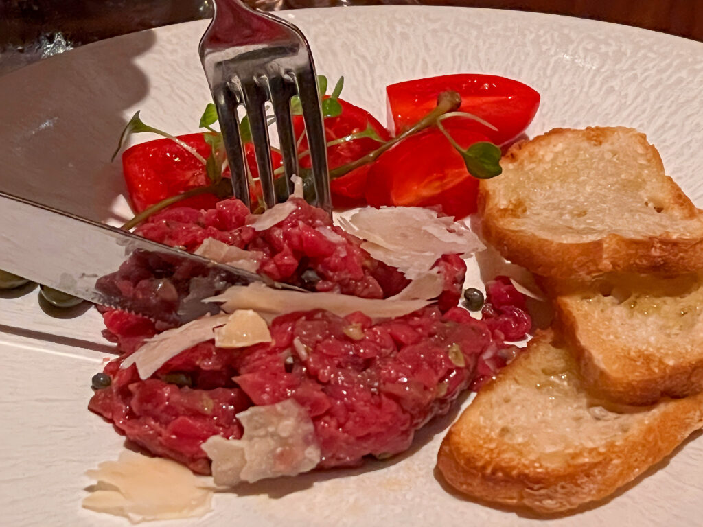 fork and knife cutting into beef tartare served with tomatoes and garlic bread