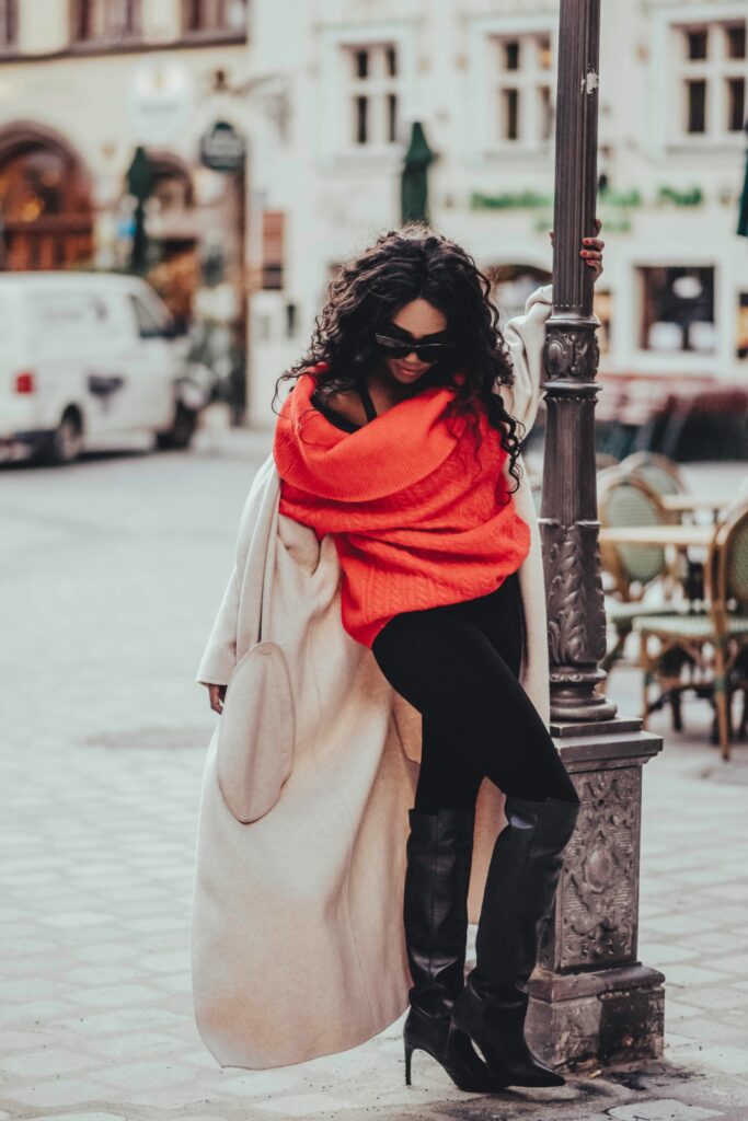 A woman showcases her minimalist style with a bold red sweater and neutral coat, leaning against a streetlamp in an urban setting.