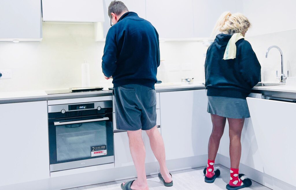 Female with kitchen towel on shoulder standing by the kitchen sink next to a man standing facing the electric stove