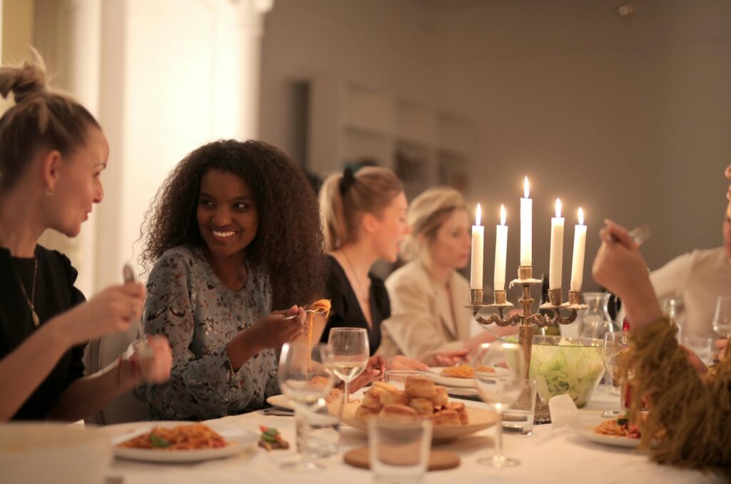 Image depicts women enjoying dinner together, eating various foods and drinking wine, while chatting and laughing