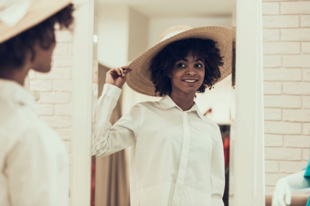 A black woman trying on a straw hat in a mirror while shopping, possibly as part of a wardrobe update. The image captures the experience of shopping alone, and reflects her search for a new look