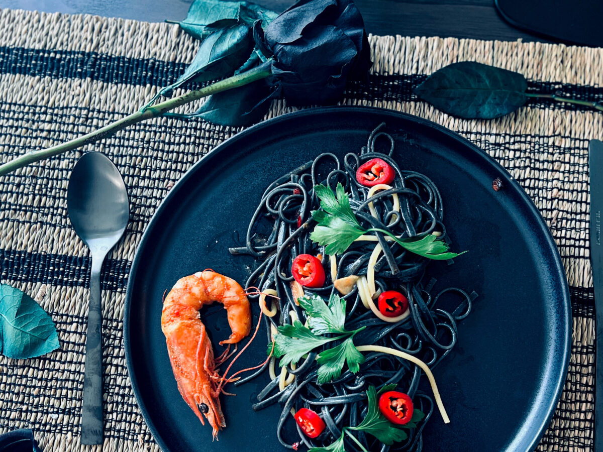 Black pasta for Halloween served on a dark plate, garnished with red chilli slices, parsley, and accompanied by a whole shrimp.