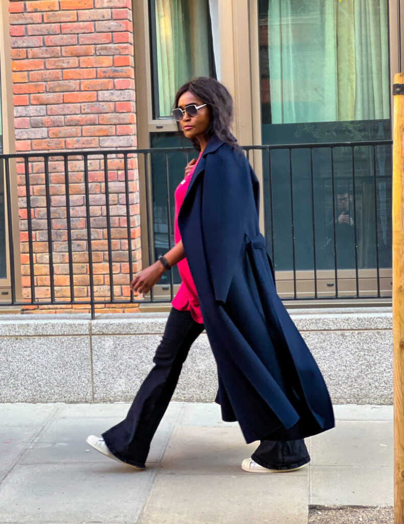 Woman wearing flared black jeans, striking pink top, white sneakers and a navy blue coat draping over her shoulder 