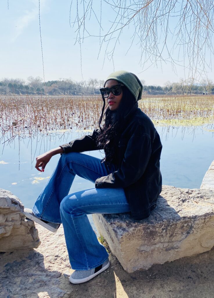Woman wearing flared blue jeans with white sneakers, black jumper, sunglasses and a green beanie sitting on a rock by a water body 