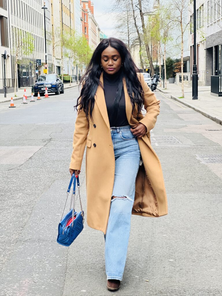 Stylish look: woman dons trendy high-waisted ripped jeans in a beautiful blue shade. Paired with a chic long brown wool blend coat for warmth and elegance. Complemented by matching brown boots and a coordinating blue handbag, creating a well-coordinated and fashionable ensemble.