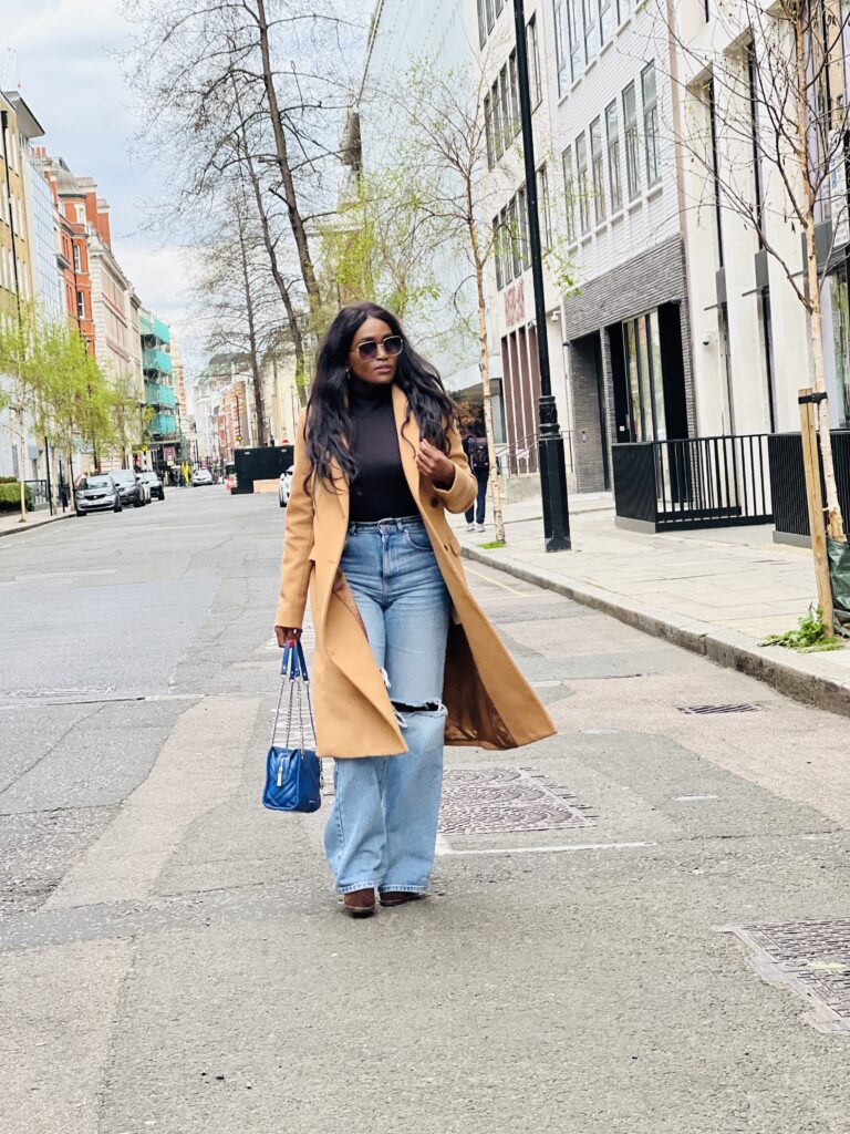 A Stylish look: woman dons trendy high-waisted ripped jeans in a beautiful blue shade. Paired with a chic long brown wool blend coat for warmth and elegance. Complemented by matching brown boots and a coordinating blue handbag, creating a well-coordinated and fashionable ensemble