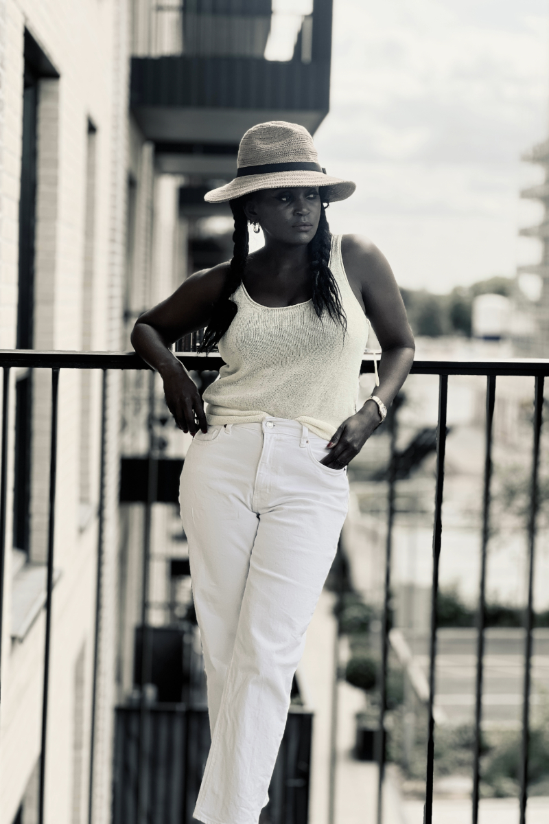 Woman wearing a straw hat, cream knit top and white trousers while standing on a balcony.