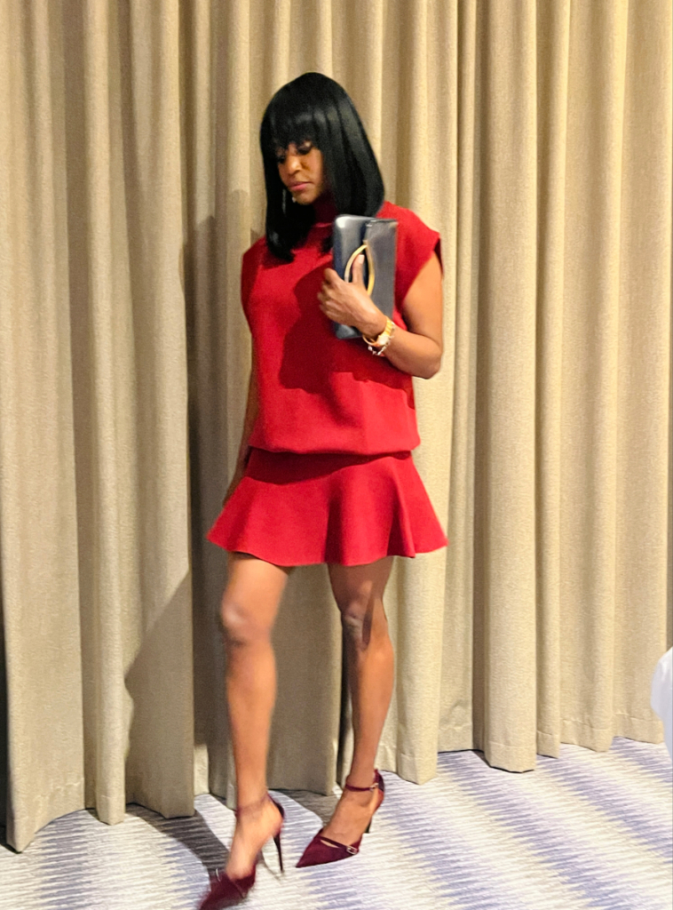Woman wearing a red sleeveless top and matching skirt with burgundy heels while holding a black clutch bag.
