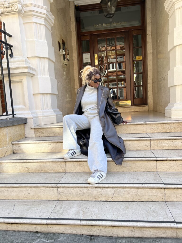 A chic black lady with blonde hair wearing a brow leather trench coat seated on stairs against a matching backdrop of a brown townhouse door.