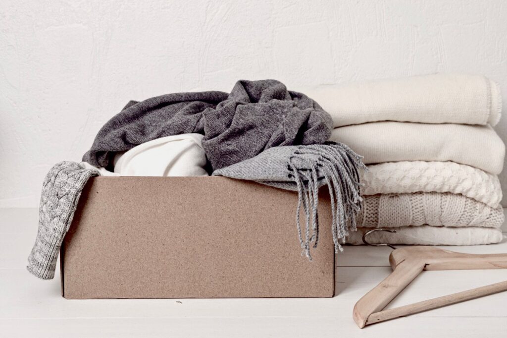 A cardboard box overflowing with winter clothing items, including cozy sweaters and soft scarves in soothing cream and elegant grey hues. Neatly folded sweaters sit beside it, along with a wooden hanger.
