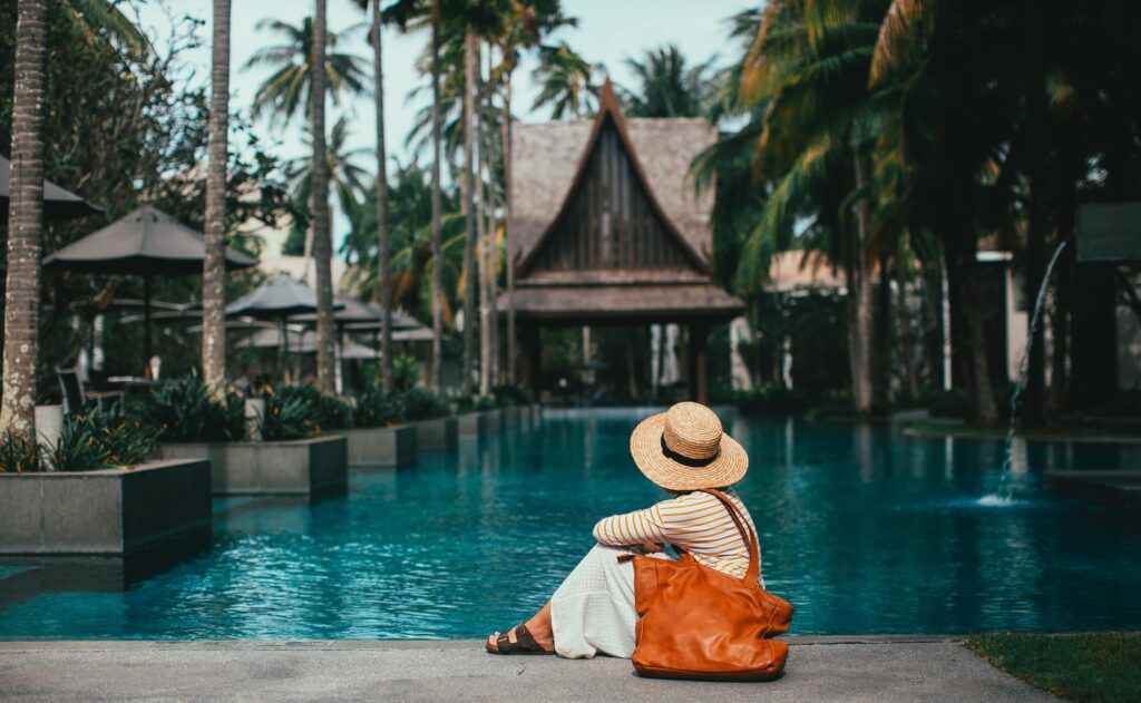 A stylish woman relaxes poolside at a resort, wearing a classic Panama hat, comfortable flat sandals, and carrying a large, vibrant orange boho bag. The tranquil scene is enhanced by the lush greenery of palm trees framing the entrance to the resort building.