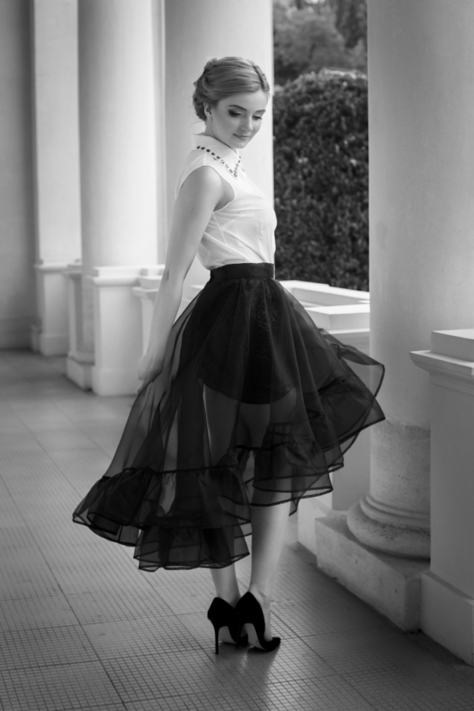 Woman wearing a white sleeveless blouse and black sheer skirt with heels standing near white columns