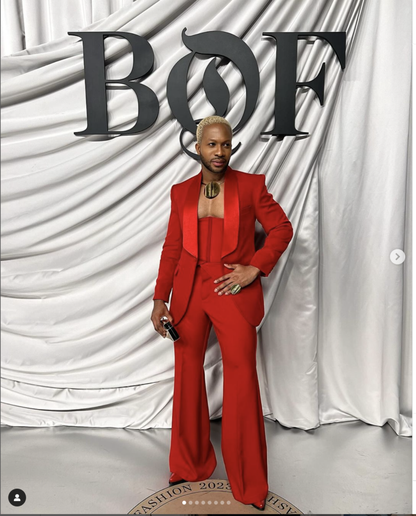 A fashion model stands in front of a draped white background with large 'BOF' letters, wearing a striking red suit. The suit features a fitted jacket and wide-legged pants. The model has short, blonde hair and wears a bold, gold necklace. This image highlights contemporary African fashion design and the model's confident pose showcases their stylish ensemble.