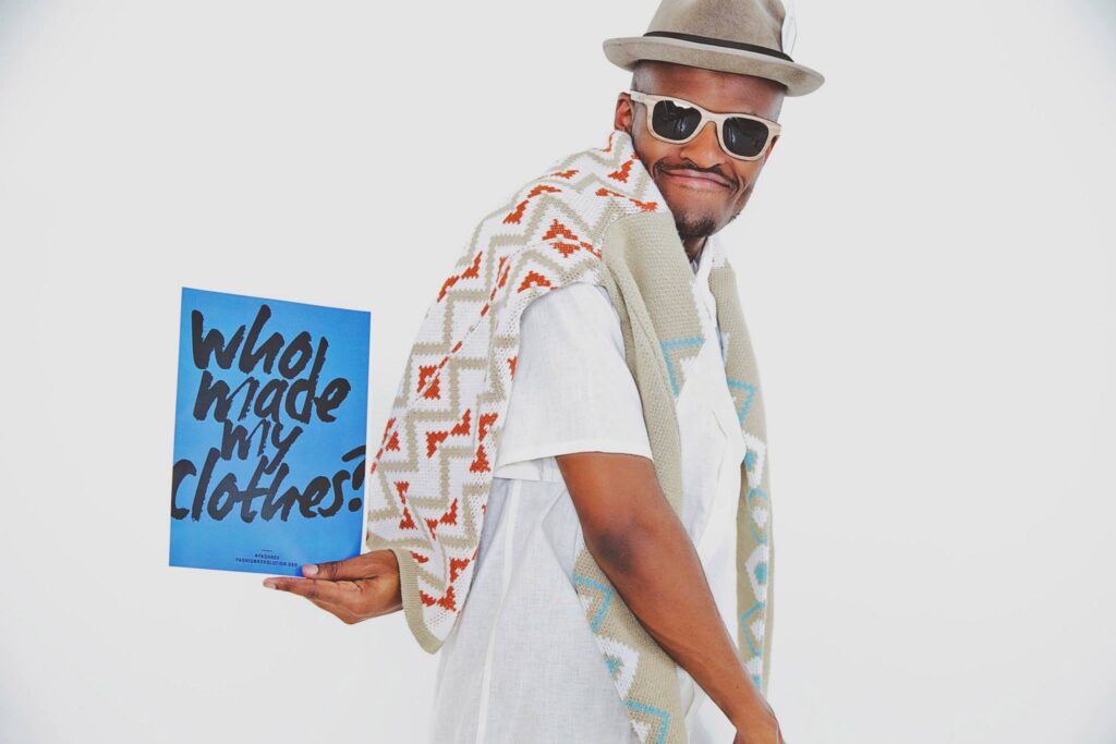 A man wearing a patterned shawl, white shirt, sunglasses, and a hat holds a blue sign that reads 'Who made my clothes?' The man smiles confidently, embodying the spirit of the fashion revolution