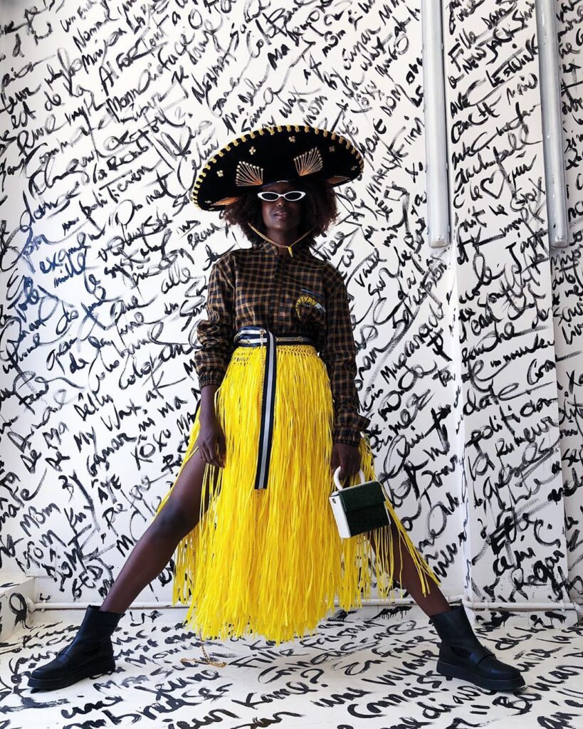 A model stands confidently in front of a graffiti-covered wall, wearing a wide-brimmed hat, checkered shirt, white sunglasses, and a vibrant yellow fringe skirt. The outfit is completed with black boots and a small handbag, showcasing the creativity of African fashion designers