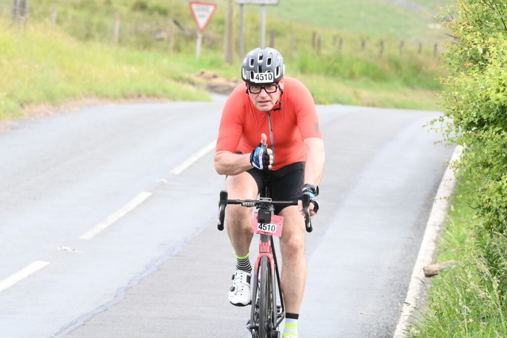 A cyclist wearing an orange jersey and helmet giving a thumbs-up while cycling on a road.