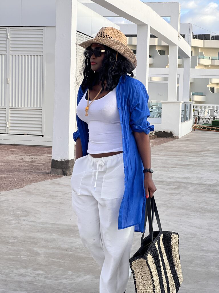 A model wearing white linen pants and a blue linen shirt from her summer capsule wardrobe accessorised with a straw bag and a straw hat.