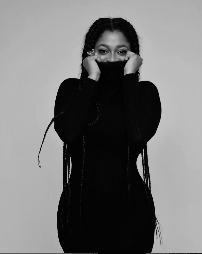 A woman with long braided hair partially covers her face with a black turtleneck sweater, highlighting her eyes in a black and white photo. This stylish portrayal reflects contemporary African fashion design