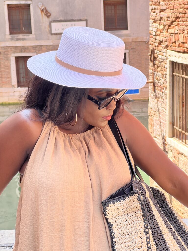 A black woman in beige top, white panama hat, and sunglasses carrying a tote bag and standing beside Venetian canal.