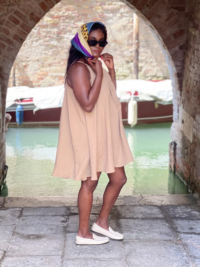 Woman in a neutral-colored sleeveless dress, accessorized with a colorful headscarf, sunglasses, and white loafers, posing under an archway by a canal in Venice, Italy.
