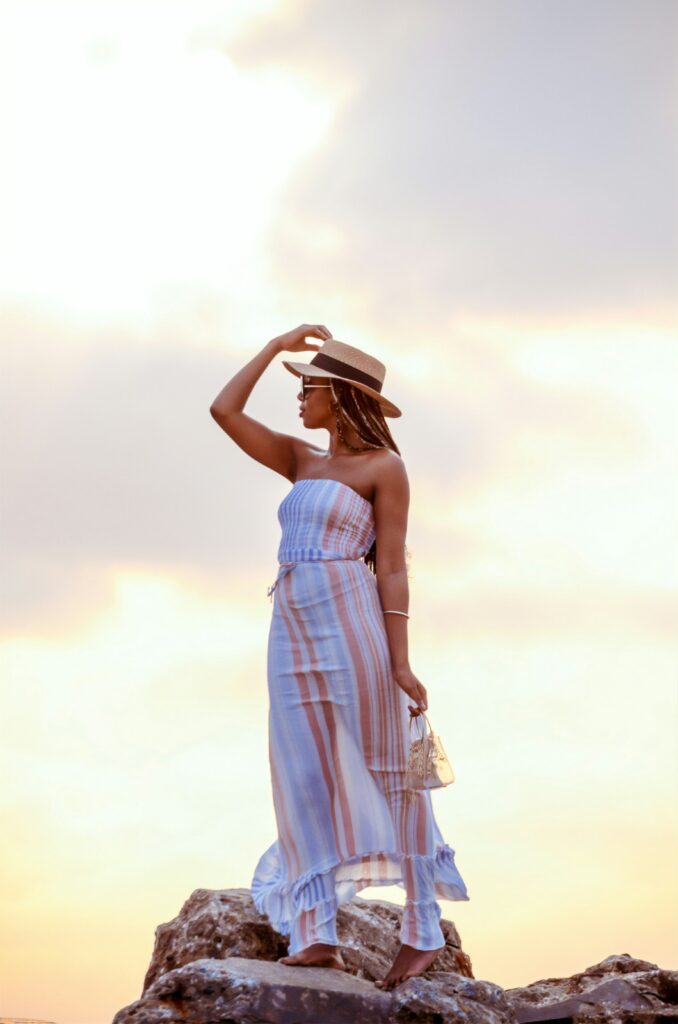 Fashionable woman in a striped sundress and sunhat, holding a clear purse on a beach at sunset.