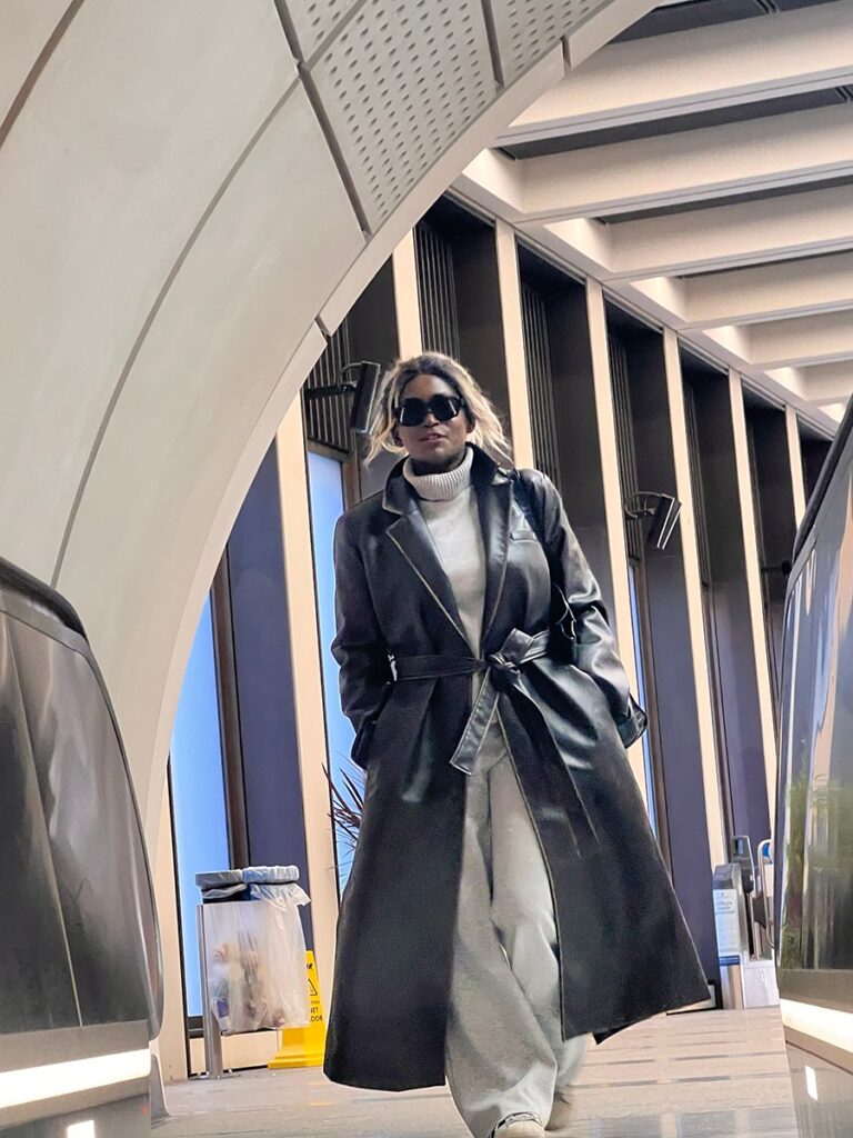 A woman showcasing how to transition your summer outfits into fall while wearing a long black leather trench coat layered over a grey oversized outfit in a modern urban setting.