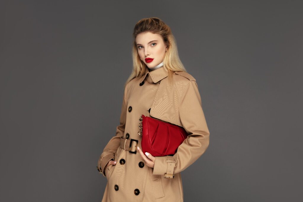 A blonde woman poses confidently in a beige trench coat, holding a red clutch bag against a grey background, exuding classic style.