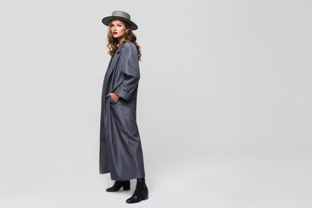A woman in a long grey coat, wearing a wide-brimmed grey hat, standing sideways with hands in pockets.
