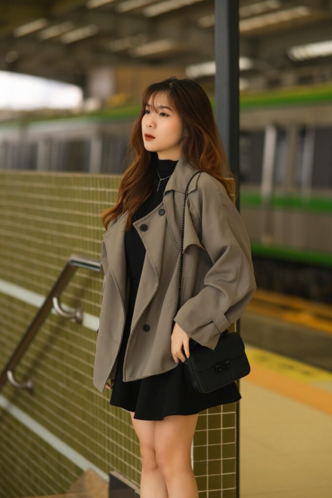 A woman in a cropped trench coat stands by a tiled wall at a train station, with a black purse slung over her shoulder.