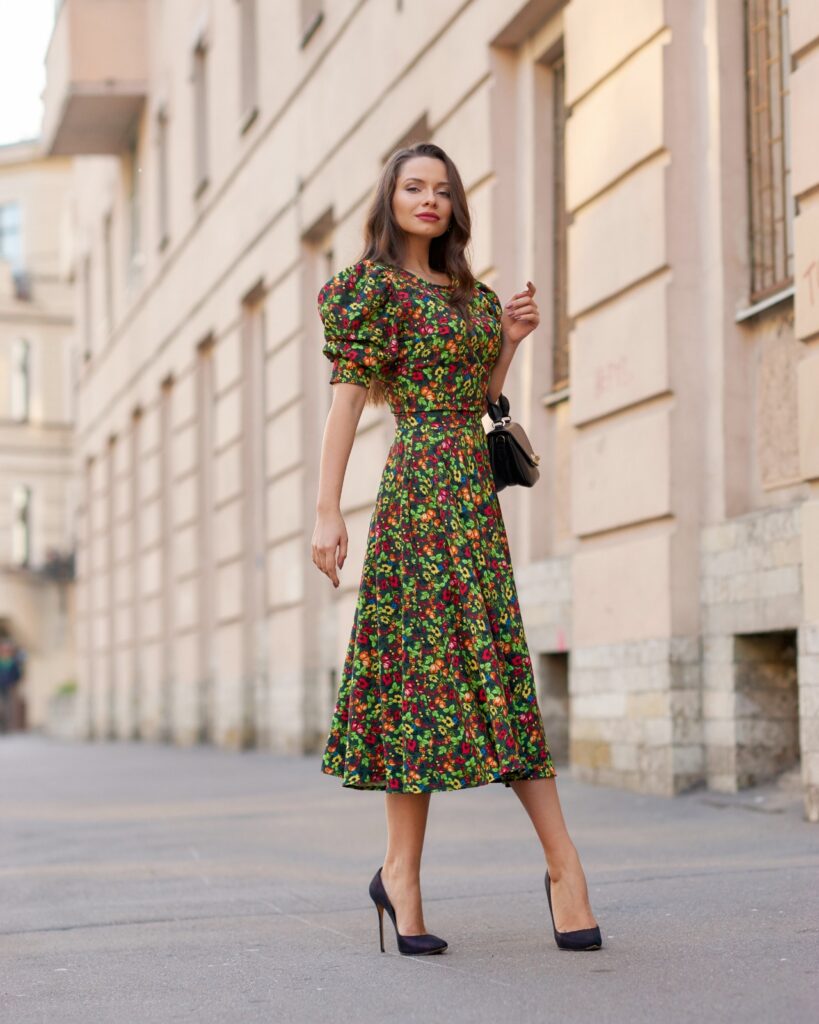 A woman is standing on a city street, wearing a floral, puff-sleeve, mid-length dress in warm tones from her fall outfits collection. In one hand she holds a small handbag that matches her black high heeled shoes.