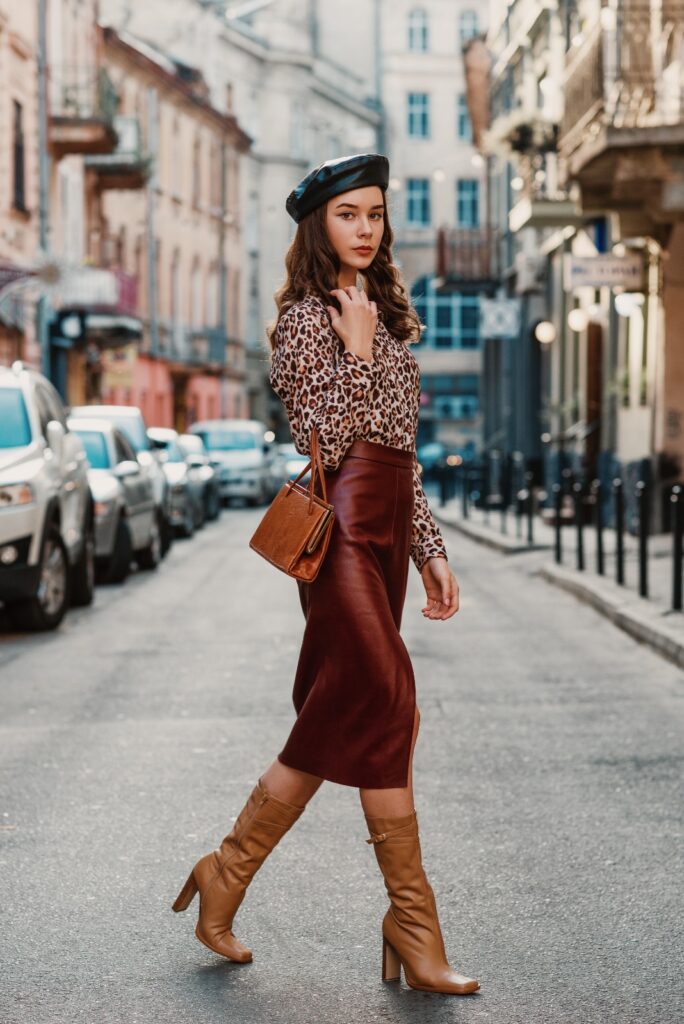 A woman in a burgundy leather skirt, leopard print blouse, slouchy brown boots, a matching bag, and a black beret walking down a city street.