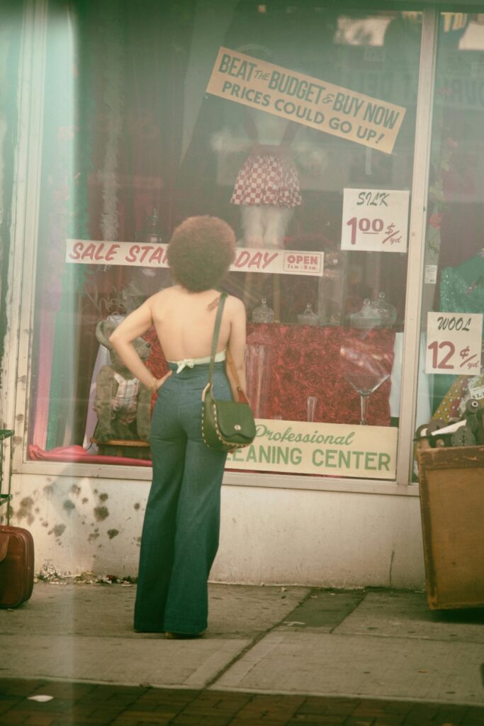  A woman with an afro hairstyle wearing flared jeans and a halter top, looking into a thrift shop window displaying sale signs.