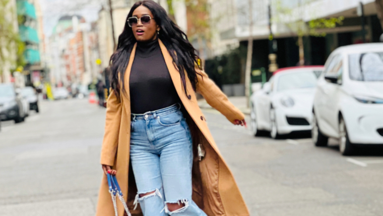 Stylish Black woman walking confidently down a city street wearing a camel coat, black turtleneck, and ripped jeans – showcasing how fashion choices like bold outerwear and fitted denim can boost mood and motivation.