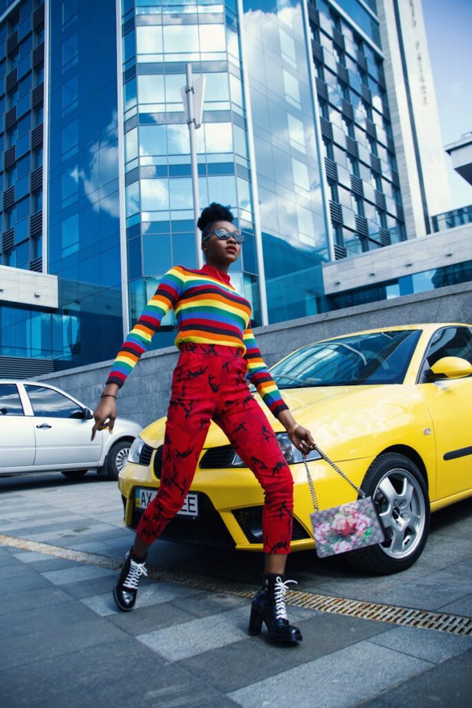 A woman in a vibrant, multicolored striped sweater and red patterned pants, walking confidently in an urban setting with modern buildings and cars in the background.