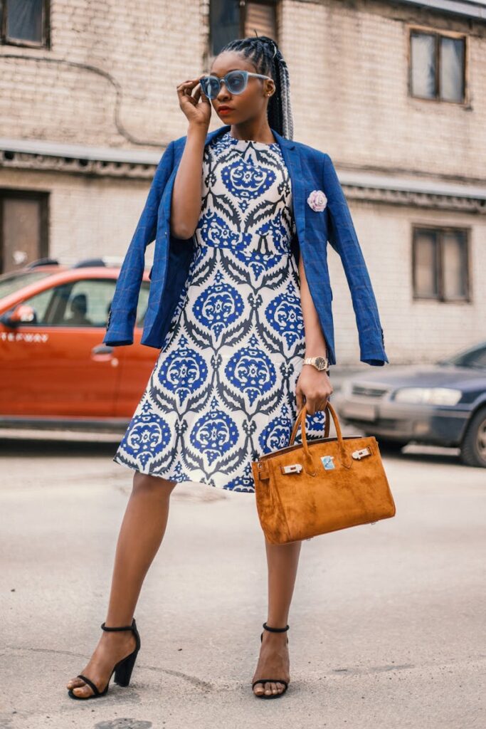 A woman in a blue and white patterned dress, complemented by a blue blazer and a brown handbag, posing confidently in an urban setting.