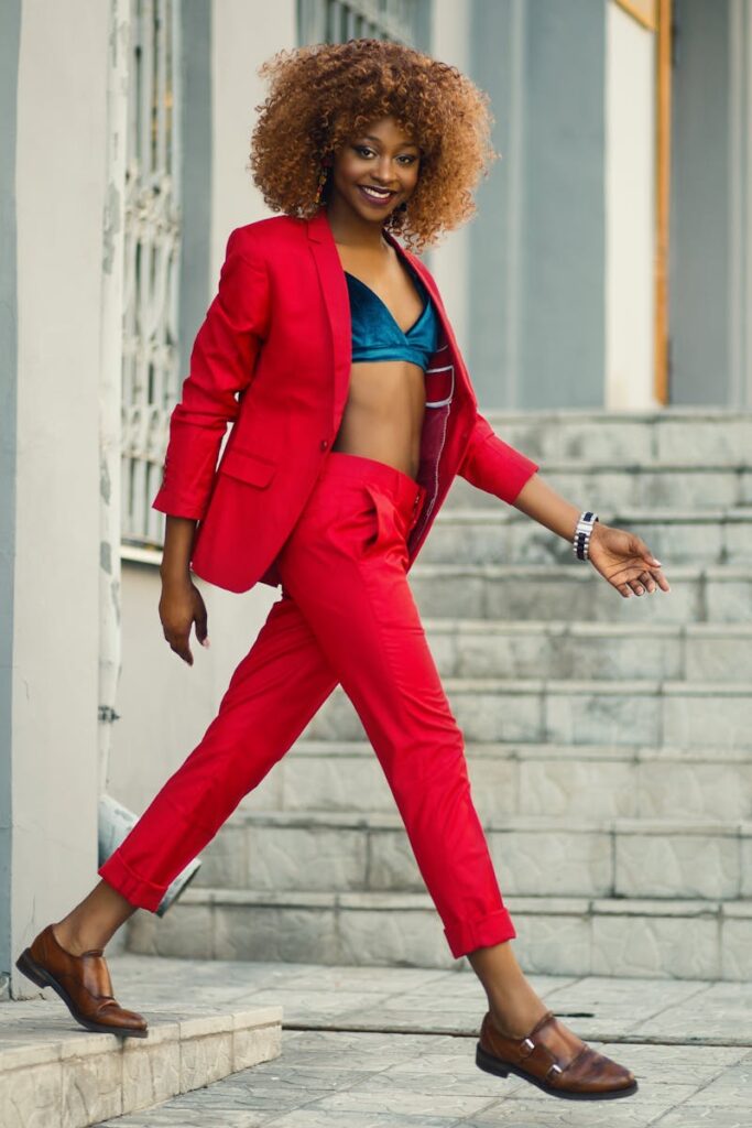 A woman in a striking red suit confidently marches across a street with a smile on her face. The suit, a bold choice from her fall outfits collection, is paired with brown Mary Jane shoes, adding a touch of classic elegance to her powerful stride.