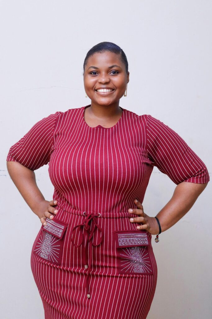A smiling woman wearing a maroon dress with white vertical stripes, posing confidently with hands on hips.
