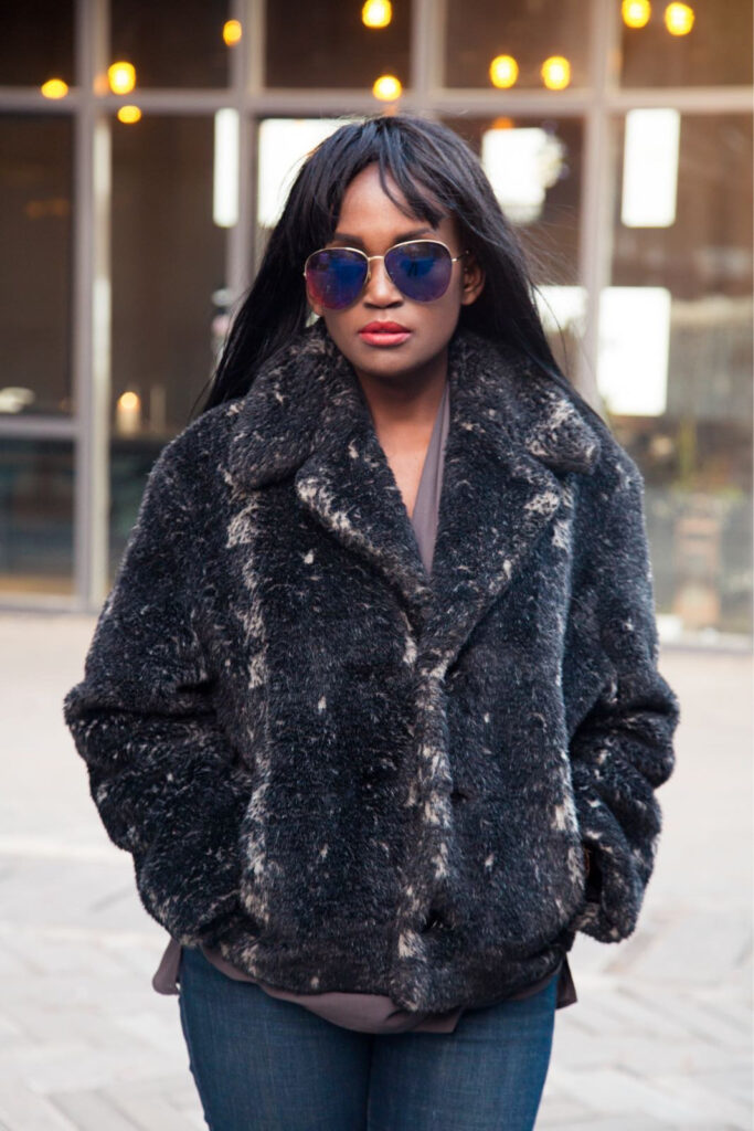 A woman in a black faux fur jacket and blue sunglasses stands outdoors, showcasing 2024 fall outfit trends.