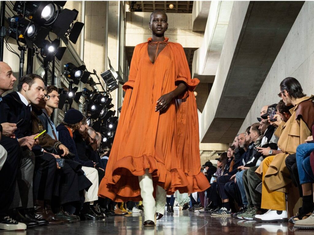 A stunning African model strides confidently down the runway wearing a voluminous orange gown. The dress features billowing sleeves and a ruffled hemline that flows gracefully with each step.