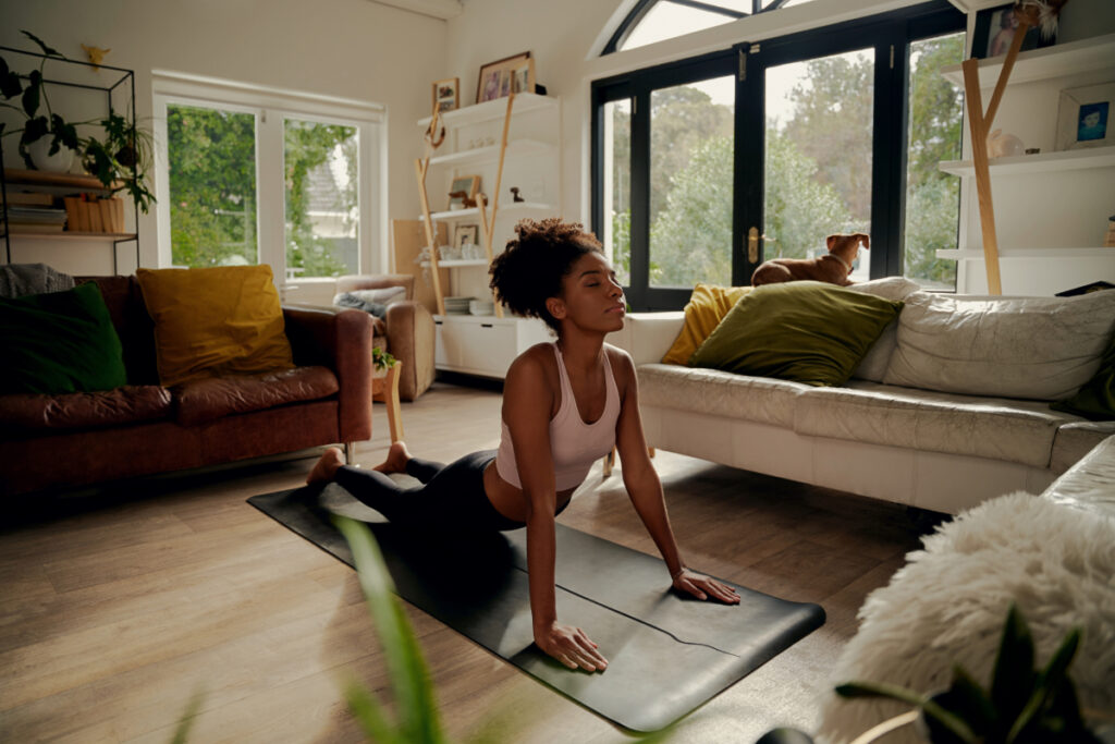 A woman practicing yoga in a bright, cosy living room, performing a cobra pose on a mat with her eyes closed, while a dog looks out of the window.