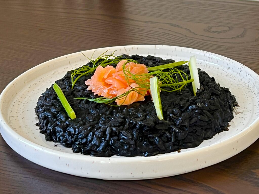 Halloween recipe: Black squid ink risotto topped with vibrant pink salmon and garnished with fennel, served on a white plate.