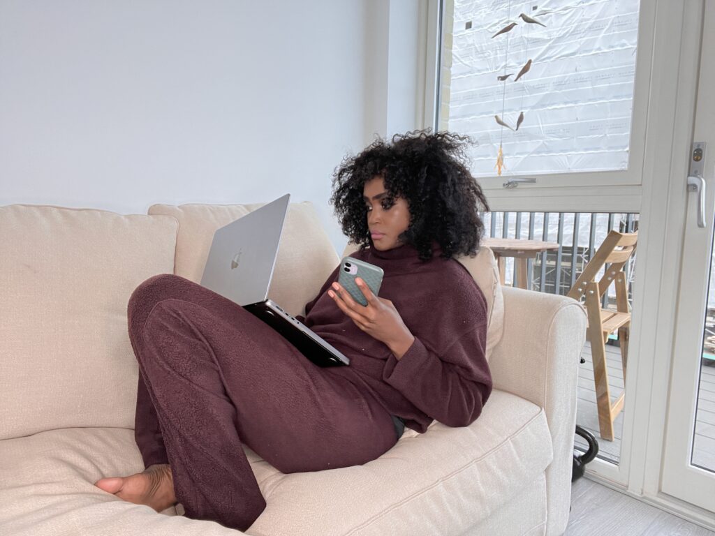 Setting boundaries with tech - Woman sitting on a beige sofa using a laptop while holding a phone, looking focused.