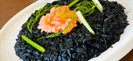 Black risotto, on a white plate, granished with smoked salmon to give a contrasting orange hue, as well as greenish vegetable stems.