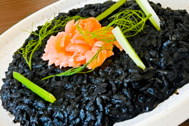 Black risotto, on a white plate, granished with smoked salmon to give a contrasting orange hue, as well as greenish vegetable stems.