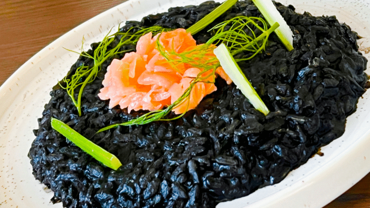 Black risotto, on a white plate, granished with smoked salmon to give a contrasting orange hue, as well as greenish vegetable stems.