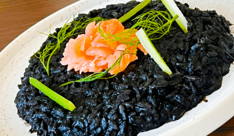 Black risotto, on a white plate, granished with smoked salmon to give a contrasting orange hue, as well as greenish vegetable stems.