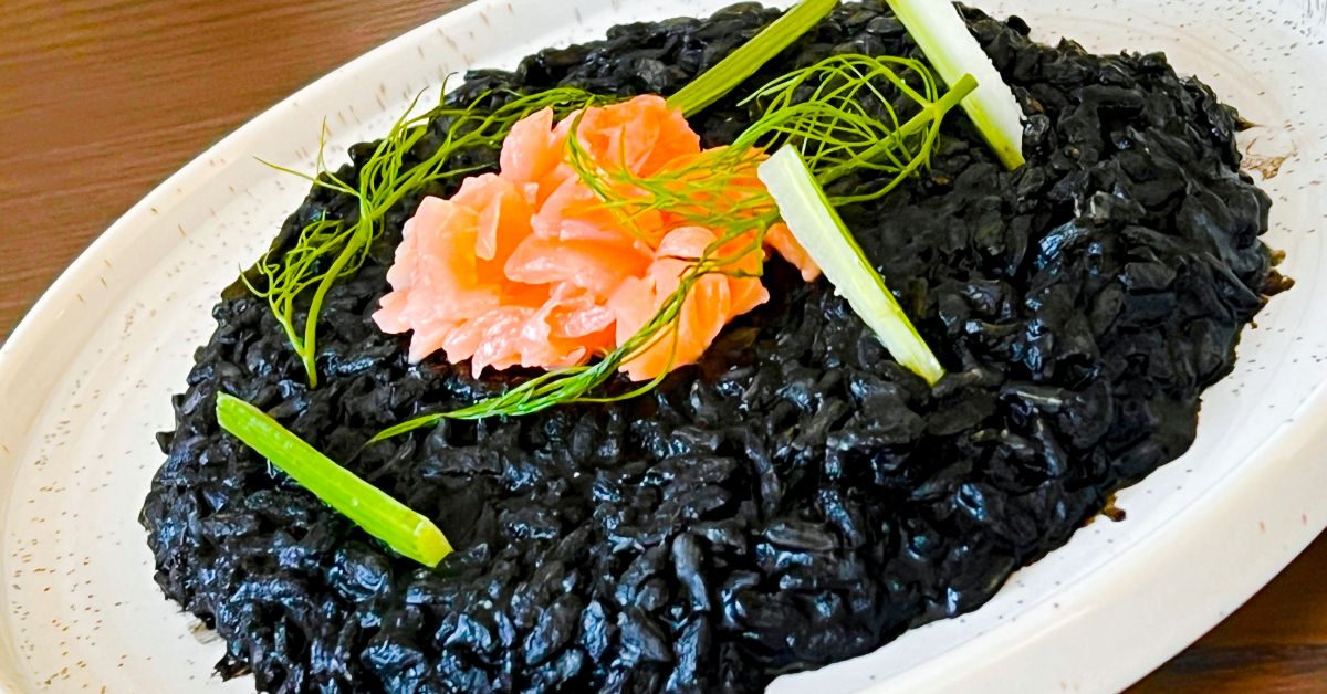 Black risotto, on a white plate, granished with smoked salmon to give a contrasting orange hue, as well as greenish vegetable stems.