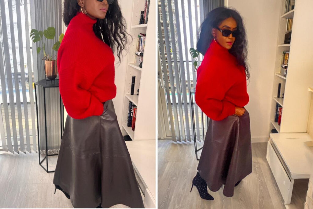 Woman in a red turtleneck sweater with a leather midi skirt and heeled boots, standing indoors near a bookshelf and plant.